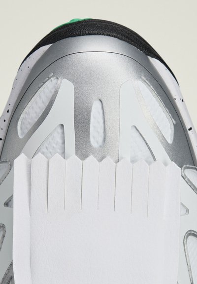Parte superior de zapatilla deportiva con una tapa de talón metálica plateada, cuerpo de malla blanca y acentos blancos texturizados con un borde dentado. Tapa verde en la parte superior.