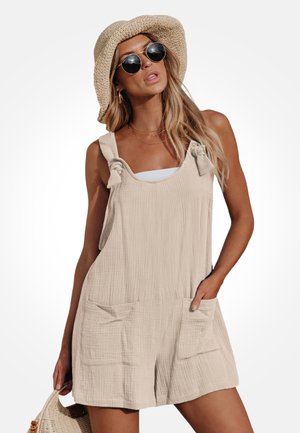 Femme portant une combinaison-short en lin beige sans manches avec poches et nœuds sur les bretelles, un chapeau de paille, des lunettes de soleil rondes et tenant un sac tissé.