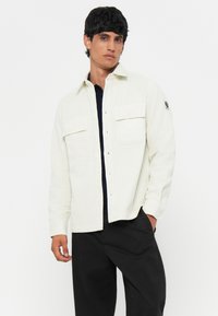 Camicia di velluto a coste bianca con due tasche frontali e chiusura a bottoni, indossata sopra una maglietta blu scuro, abbinata a pantaloni neri.