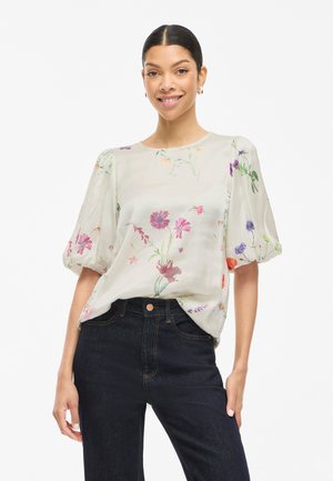 Femme portant un chemisier blanc avec un imprimé floral coloré et des manches courtes bouffantes, associé à un jean taille haute sombre, souriante.