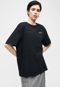 Νέος με κοντά σκούρα μαλλιά, που φοράει μια μαύρη φαρδιά μπλούζα Columbia και ανοιχτό γκρι παντελόνι cargo, στέκεται με τα χέρια στη μέση.