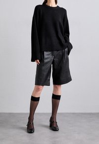 Musta jättimallinen neulepusero, jossa resorireuna, nahkashortsit, ohuet mustat polven yli ulottuvat sukat, joissa tukevat raidat, ja mustat Mary Jane -malliset korkokengät.