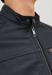 Närbild av en person som bär en svart texturerad läderjacka med silverdragkedja och tryckknappskrage mot en neutral bakgrund.