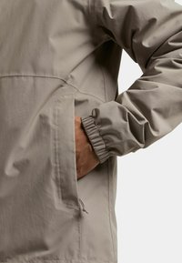 Chaqueta ligera de color gris claro con puños elásticos, un bolsillo lateral y un detalle de cordón en el dobladillo para ajuste regulable.