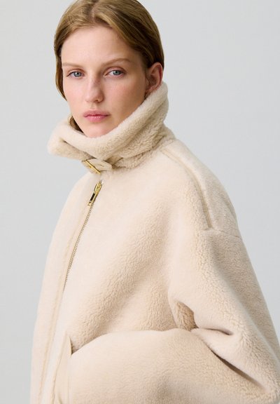 Giacca in shearling beige con colletto alto, chiusura a cerniera e tasche laterali; texture morbida con una silhouette rilassata e dettagli in oro.