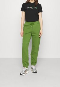 Černé tričko s krátkým rukávem s logem "CALVIN KLEIN JEANS", spárované s volně padnoucími zelenými tepláky a st říbrnými sportovními botami.