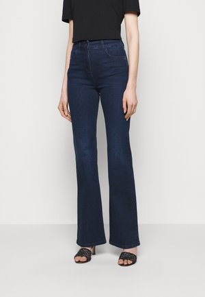 Person stående iført mørkeblå højtaljede vide jeans, sort kortærmet top og sorte åbne hæle på en ensartet lys baggrund.