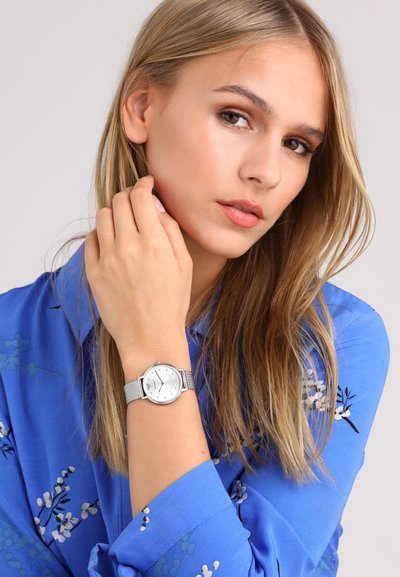 Junge Frau mit langen blonden Haaren, die ein blaues Blumenhemd trägt, berührt ihr Gesicht und zeigt eine silberne Armbanduhr mit weißem Zifferblatt.