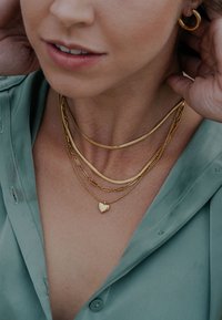 Zlatni ogrlice u slojevima s privjeskom u obliku srca, različitih stilova lanaca i glatkom teksturom, nošene preko svijetlozelene bluze.