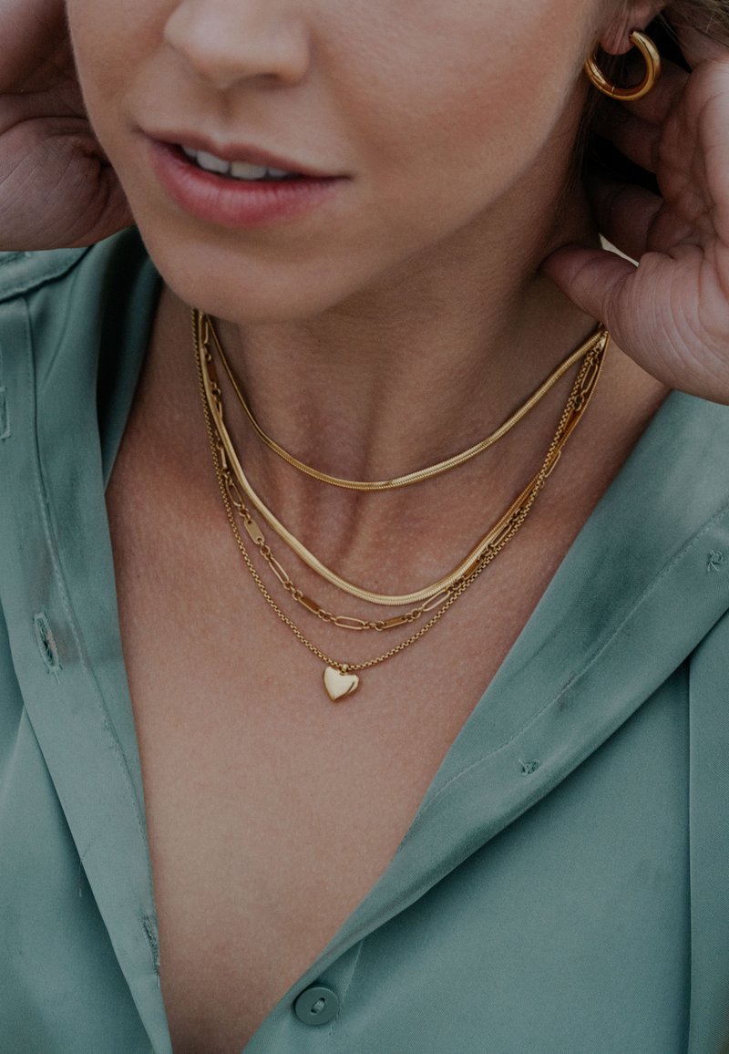 Zlatni ogrlice u slojevima s privjeskom u obliku srca, različitih stilova lanaca i glatkom teksturom, nošene preko svijetlozelene bluze.