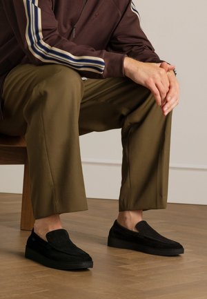 Chaussures noires en daim à enfiler avec une surface texturée et une semelle en caoutchouc plate, associées à un pantalon large vert olive et à une veste marron zippée.
