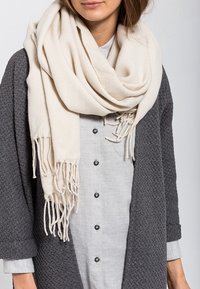 Femme portant une grande écharpe crème à franges, un manteau gris texturé et une chemise à boutons gris clair.
