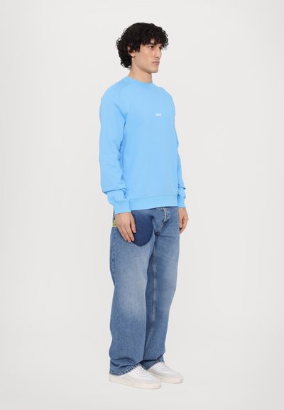 Sudadera azul claro con puños y dobladillo acanalados; presenta un pequeño logo blanco en el pecho. Se lleva con unos pantalones vaqueros de denim azul de corte suelto y zapatillas deportivas blancas.