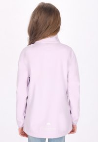 Chaqueta impermeable de color morado claro, con un alto cuello, mangas largas y un logo circular cerca del dobladillo. Textura suave y elementos de diseño mínimos.