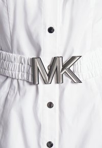 Vêtement blanc avec boutons-pression noirs et taille élastique avec une grande boucle de ceinture métallique argentée avec le logo "MK".