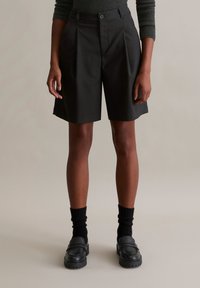 Zwarte op maat gemaakte shorts met plooien, voorzien van een knoopsluiting, zijzakken en een soepele, lichte stof. G gedragen met zwarte kniekousen en instappers.