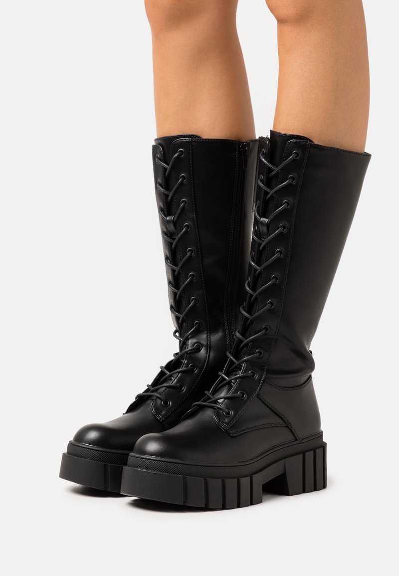 Bottes en cuir noires montantes jusqu'aux genoux avec des semelles épaisses rainurées, des lacets à l'avant et des fermetures éclair sur les côtés, portées par une personne debout sur un fond blanc.