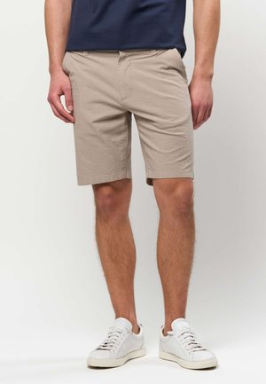 Mężczyzna w beżowych, teksturowanych spodenkach do kolan, granatowej koszuli i białych trampkach, stojący na jednolitym tle.