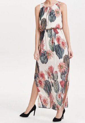 Femme portant une robe maxi blanche sans manches avec un imprimé coloré de cactus et de fleurs, associée à des chaussures à talons noirs.