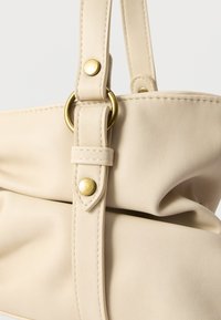 Sac à main en cuir synthétique beige avec un design arrondi, une surface texturée, des ferrures en laiton et une poignée supérieure avec des détails de couture.