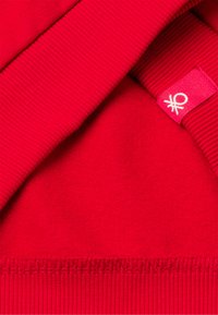 Primo piano di un tessuto di maglia rosso con cuciture visibili e una piccola etichetta rossa con un logo circolare bianco.