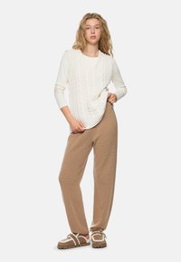 Young woman wearing a white cable-knit sleeveless sweater, cream long-sleeve shirt, beige knit pants, and shearling-lined slippers.