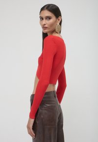 Frau mit langen dunklen Haaren trägt einen roten gerippten kurzen Pullover, große goldene strukturierte Ohrringe und hoch taillierte braune Lederhosen.