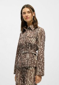Braune gemusterte Jacke mit langen Ärmeln, Fronttaschen und einem Gürtel in der Taille. Der Stoff hat ein strukturiertes Design mit helleren Akzenten.