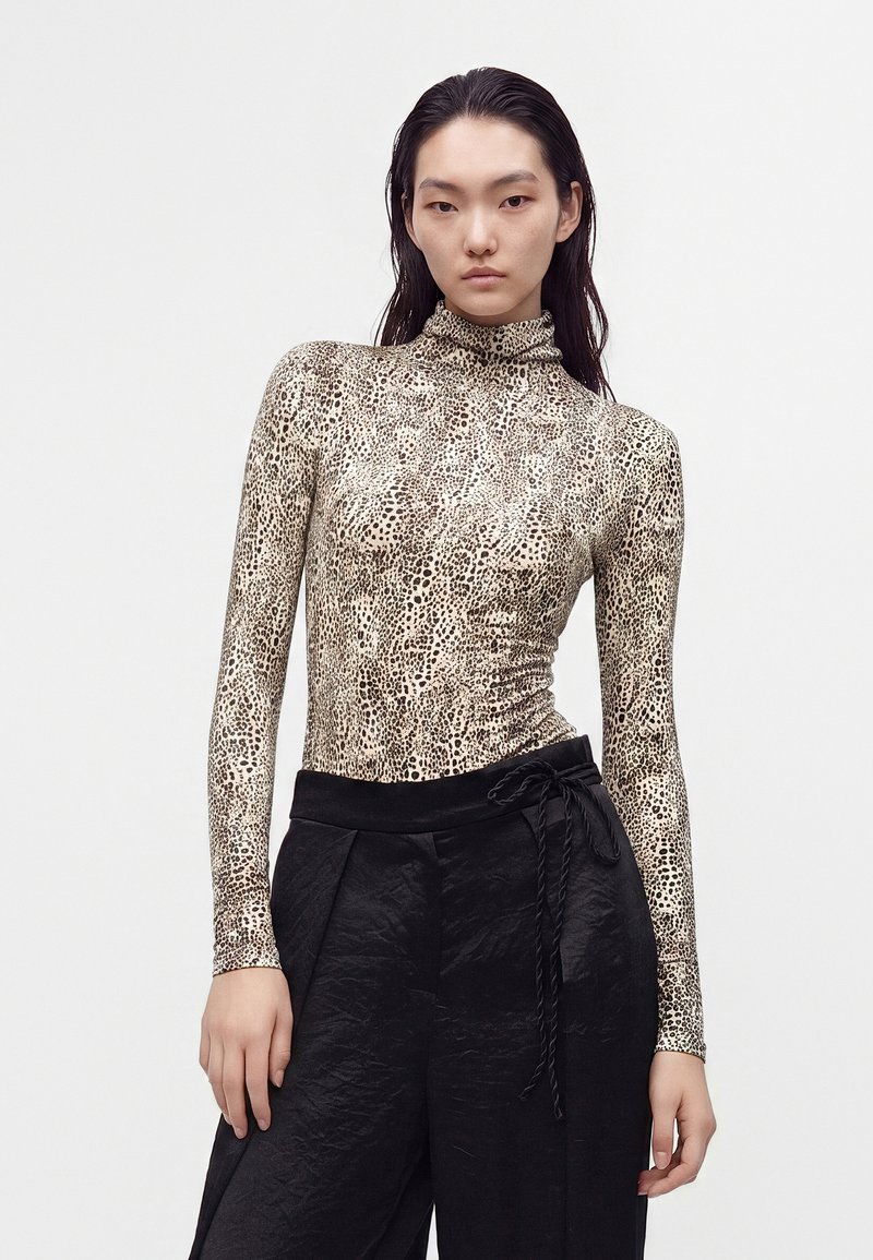 Femme aux longs cheveux foncés portant un haut moulant à manches longues imprimé léopard et un pantalon noir taille haute avec une ceinture nouée.