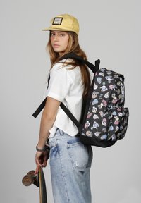 Mochila negra con patrones coloridos de temática skate, correas ajustables y forma redondeada. Llevada por una persona que sostiene una patineta.