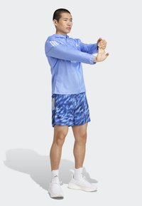 Veste zippée bleu clair à manches longues, associée à un short bleu tie-dye. Le modèle porte des chaussures de sport blanches et des chaussettes, en train de réaliser un étirement.