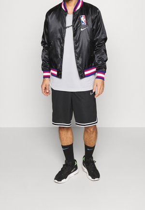 Chaleco varsity de satén negro con puños a rayas en rojo, blanco y azul. Camiseta de cuello redondo gris, pantalones cortos deportivos negros y zapatillas negras con acentos verdes.