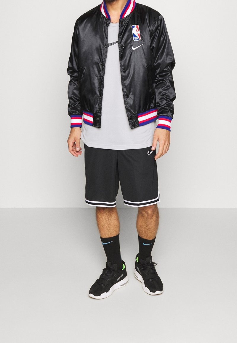 Giacca varsity in satin nera con polsini a strisce rosse, bianche e blu. Maglietta a girocollo grigia, pantaloni sportivi neri e sneaker nere con dettagli verdi.