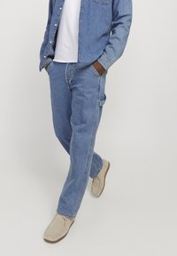 Vaqueros de mezclilla azul claro con un corte relajado, combinados con una camisa de mezclilla de botones y zapatos de lona beige. Fondo neutro.