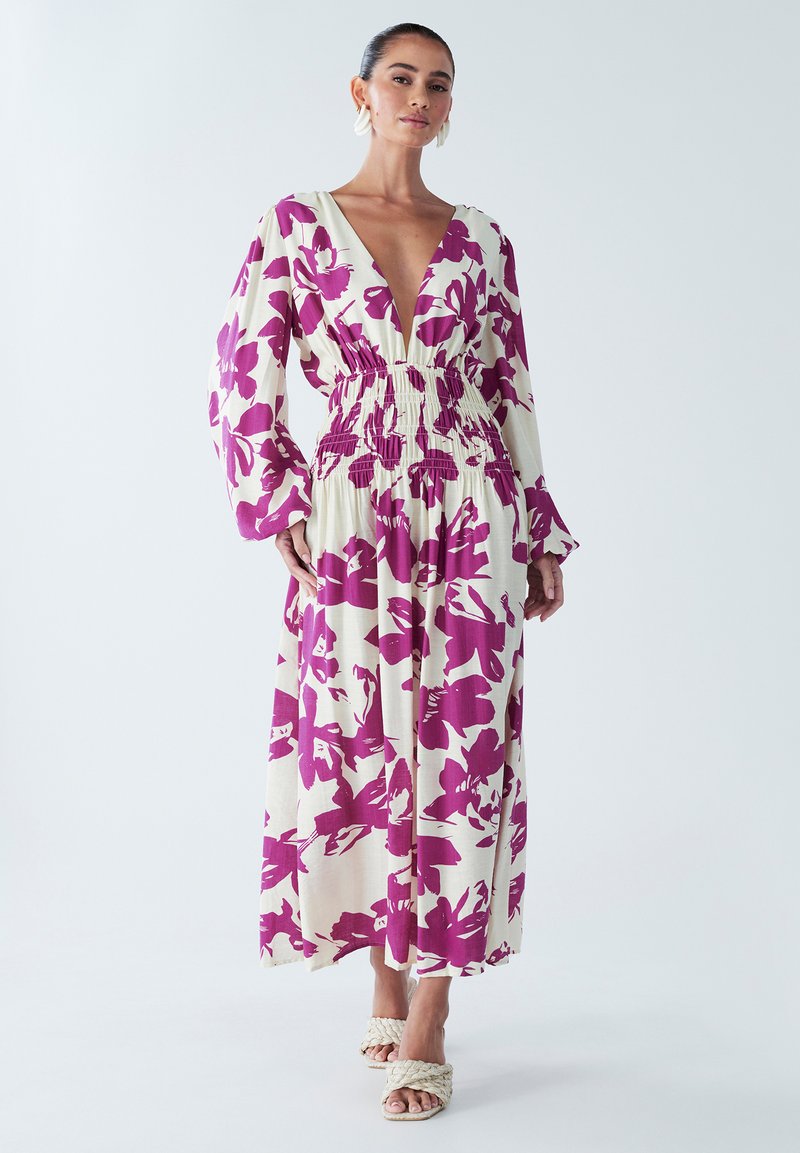 Robe maxi florale avec un décolleté en V profond, des manches longues et une taille élastique. Présente de grandes fleurs violettes sur un fond crème. Portée avec des sandales tressées.