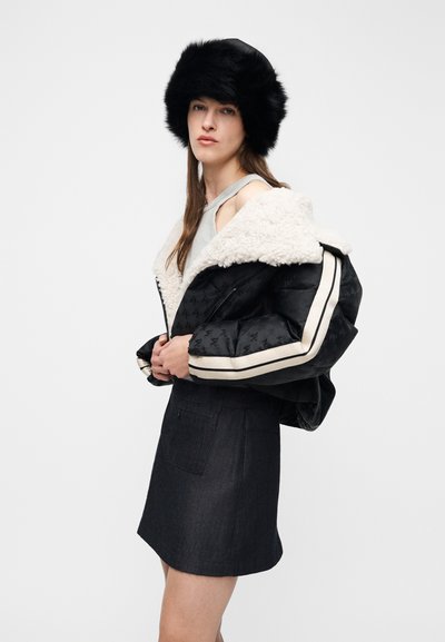 Γυναίκα που φοράει μαύρο γούνινο καπέλο, μαύρο μπουφάν με λευκές ρίγες και γιακά από shearling πάνω από γκρι αμάνικη μπλούζα και μαύρη φούστα, ποζάροντας πλαγίως.