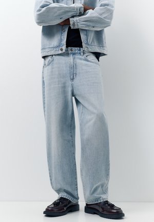 Person iført lysblå denimjakke og jeans med vide ben samt sorte loafers med kvaster, stående med armene krydset mod en hvid baggrund.