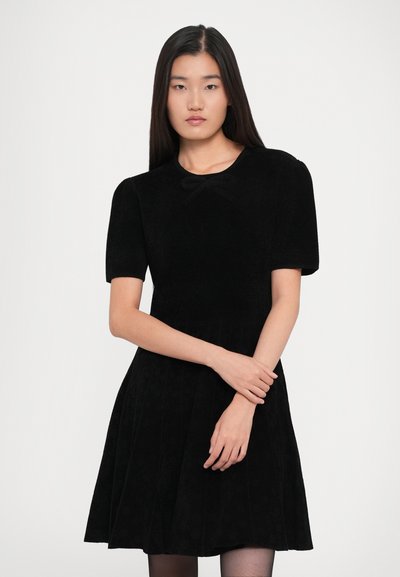 Woman with long straight black hair wearing a short-sleeve black dress with a small bow at the neckline, standing against a plain background.