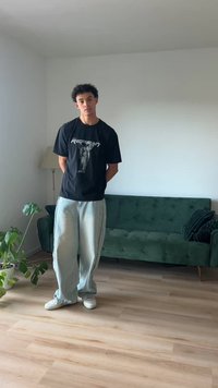 Juodas marškinėliai su grafikiniu spausdiniu, laisvi šviesiai mėlyni džinsai, balti batai, stovi patalpoje šalia žalios sofos ir augalo.