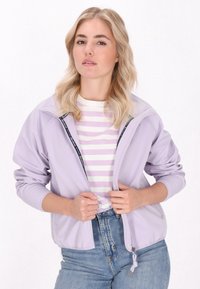 Svetlo lilav zip-up jopič s stoječim ovratnikom, ki ima stranske žepe in mehak material. Obut nad črtasto srajco in modrimi kavbojkami.
