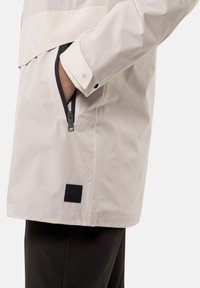 Lichte crème jacket met een ritszak, drukknopen bij de mouwen en een zwart merkembleem aan de onderkant. Kenmerkt zich door een soepele textuur en ademend ontwerp.