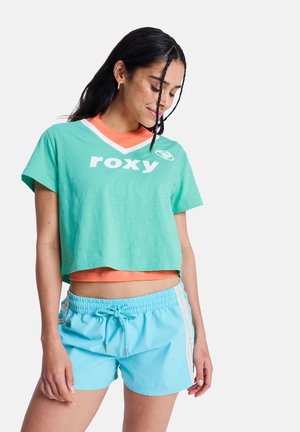 Vrouw met lang donker haar, een teal cropped T-shirt over een oranje top en lichtblauwe shorts met zijstrepen, staand tegen een witte achtergrond.