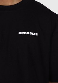 Mustast puuvillane T-särk, millel on valge "DROPSIZE" logo julges, plokikujulises fondis vasakul rinnapiiril. Lühikesed varrukad.