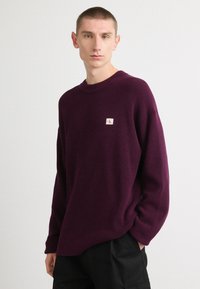 Purple knit sweater with a crew neckline and ribbed texture. Features a small logo patch on the left chest. Loose-fit with dropped shoulders.