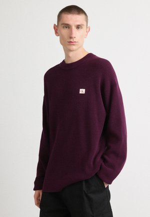 Lila Strickpullover mit Rundhalsausschnitt und gerippter Textur. Verfügt über ein kleines Logo-Patch auf der linken Brust. Lockere Passform mit überschnittenen Schultern.