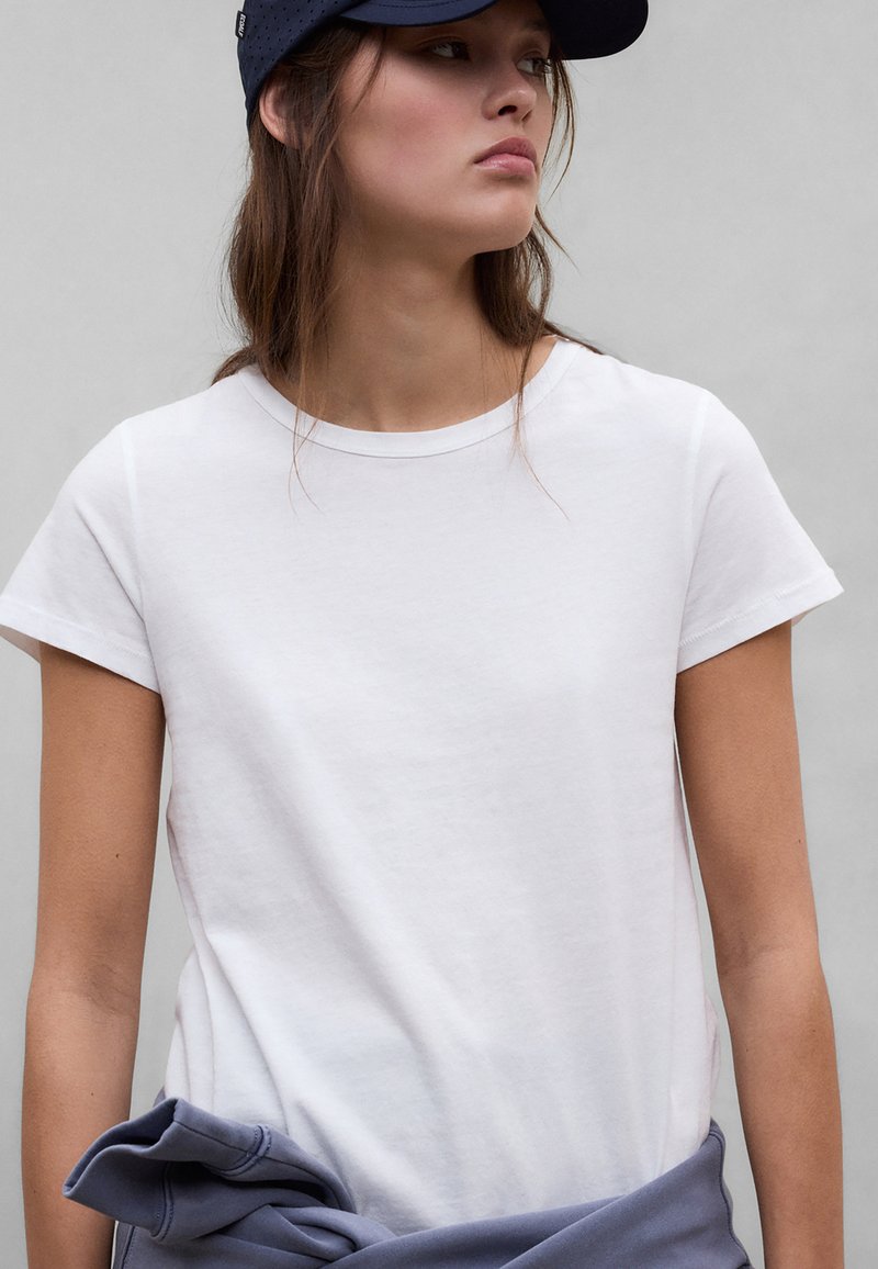 Νέα γυναίκα που φοράει ένα απλό λευκό t-shirt και μπλε καπέλο, με ένα μπλε φούτερ δεμένο γύρω από τη μέση της, κοιτάζει στο πλάι.