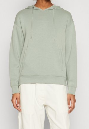 Osoba ubrana w jasnozieloną bluzę z kapturem i sznurkami oraz białe spodnie, stojąca na jednolitym tle.