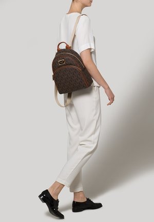 Sac à dos marron à motifs avec des accents en cuir, de forme ronde, doté de bretelles réglables et d'une poignée supérieure, porté par un modèle sur fond neutre.