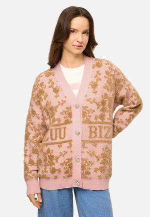 Vrouw draagt een roze en bruine bloemencardigan met knopen, een wit topje en blauwe jeans, staand met één hand op haar heup.