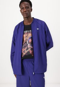 Veste de sport violette avec fermeture éclair à l'avant et poignets côtelés, portée sur un t-shirt graphique noir. Un pantalon violet assorti complète la tenue.